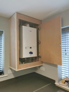 An unpainted wooden kitchen cabinet with the door open to reveal a boiler